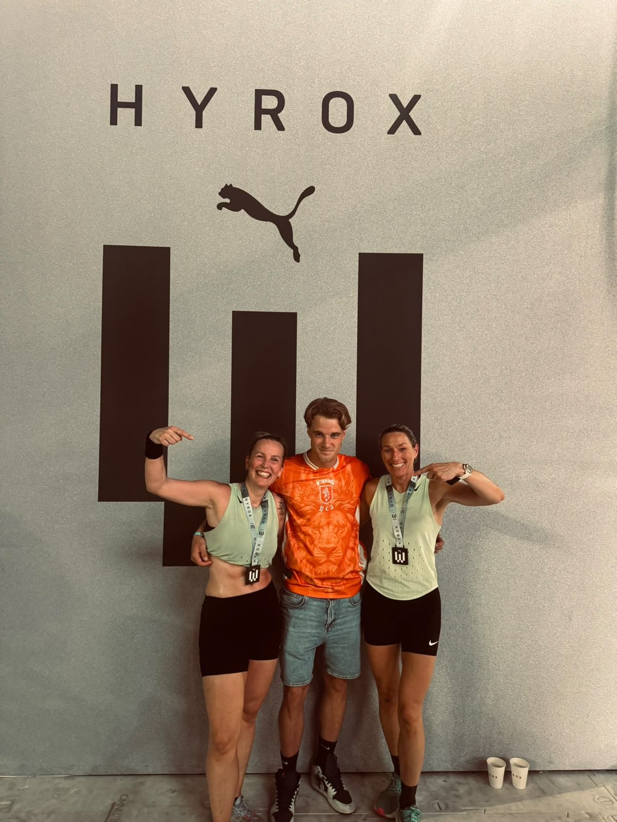 Teun met twee atleten die hun finishermedaille dragen, voor de wand van HYROX.